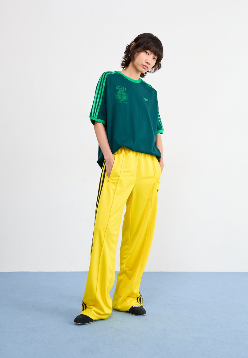 Groene katoenen T-shirt met groene strepen, voorzien van een logo; gele polyester broek met zwarte zijkanten, gedragen met zwarte instappers.