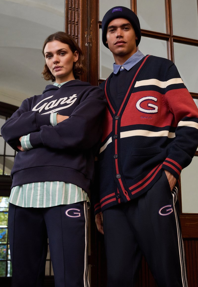 Hoodie navy avec logo "Gant" blanc, chemise rayée verte en dessous. Homme en cardigan navy à accents rouges avec rayures blanches et logo. Pantalons sombres.