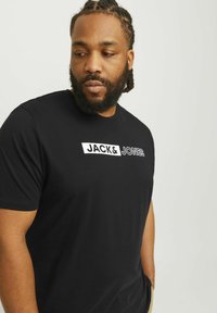 Crna majica kratkih rukava od pamuka, s bijelim logotipom "JACK & JONES" u debelom tisku preko prsa. Glatka tekstura.