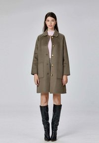 Manteau en laine vert olive avec un col, des boutons dorés et des poches avant, associé à des bottes noires brillantes jusqu'aux genoux. Design simple, mi-long.