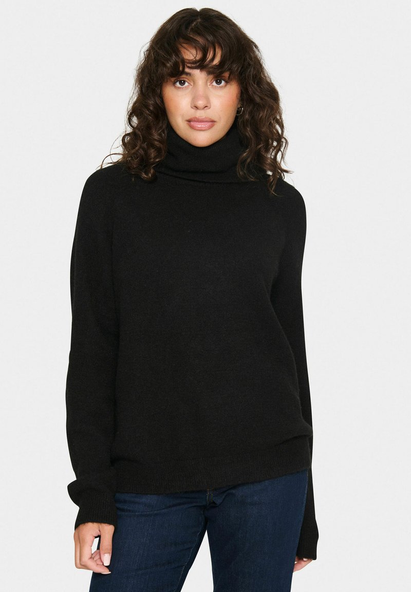 Melna turtlenecks džemperis, izgatavots no mīksta auduma, ar garām piedurknēm, brīva piegriezuma un ribotām aprocēm. Apvienots ar tumši zilām džinsām.