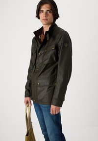 Veste imperméable vert foncé avec plusieurs poches, accents en métal doré et un col haut. Associée à un jean bleu et un grand sac en toile clair.