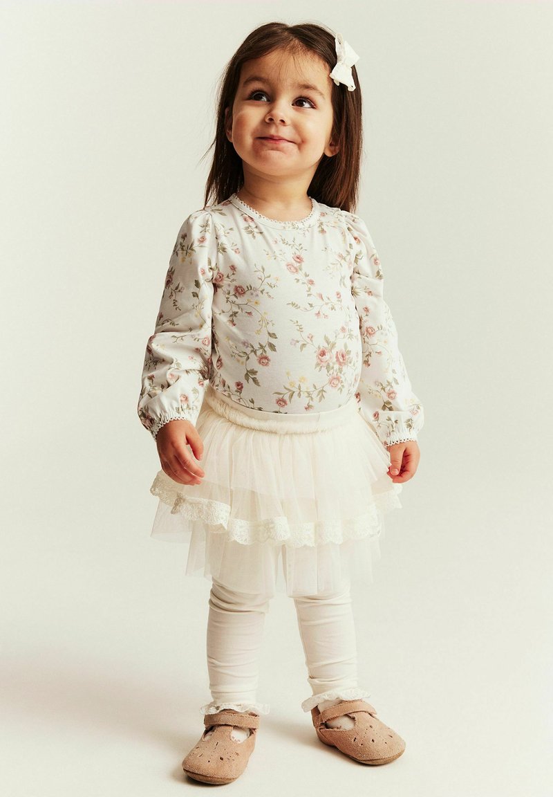 Lille pige smilende, iført blomstret langærmet top, hvid tutu-nederdel, leggings, beige sko og hvid sløjfe i håret, stående på lys baggrund.