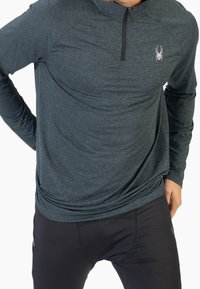 Maglietta grigia scura a maniche lunghe con mezza zip, materiale testurizzato e logo Spider sul lato sinistro del petto. Abbinata a pantaloni neri aderenti.