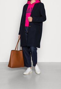 Cappotto navy su un hoodie rosa, abbinato a jeans scuri e sneakers bianche. Tiene una borsa a tracolla in pelle marrone. Texture morbide e colori a contrasto.