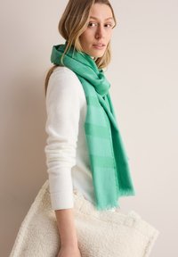 Sciarpa verde in morbido tessuto, con un delicato motivo a quadri, lasciata cadere intorno al collo; indossata con un maglione bianco e una borsa di tessuto panna.