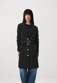 Sort bæltestøvle lavet af et struktureret stof. Har en spids krave, frontlommer og sølvknapper. Bæres over denim.