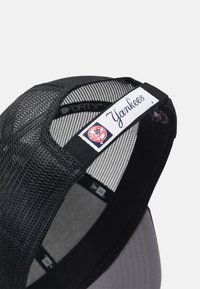 Schwarze Netzmütze mit grauem Schild, ausgestattet mit einem gestickten "Yankees"-Label und Logo auf der Rückseite, genähten Details und einem verstellbaren Riemen.