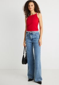 Νεαρή γυναίκα με σγουρά μαλλιά, φοράει κόκκινο ασύμμετρο αμάνικο τοπ, φαρδιά μπλε τζιν παντελόνια, μαύρα μυτερά παπούτσια, κρατώντας μαύρη τσάντα χειρός.