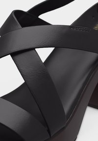 Μαύρο δερμάτινο σανδάλι με κοίλο τακούνι, φαρδιές λουρίδες σε σχήμα σταυρού και ανοιχτή μύτη, εμφανίζεται σε κοντινή λεπτομέρεια.