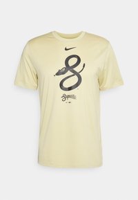 Camiseta Nike en amarillo claro, con un gráfico de serpiente negra y texto "Serpientes". Mangas cortas y cuello redondo. Textura suave.