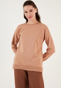 Maglia beige a maniche lunghe realizzata in tessuto morbido, con scollatura rotonda e vestibilità rilassata, abbinata a pantaloni marroni. Texture liscia, senza motivi visibili.