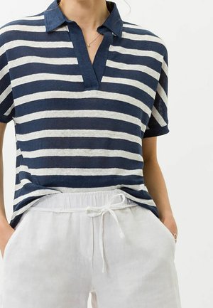 Kobieta ubrana w granatową i białą w paski koszulkę polo z krótkim rękawem, włożoną w białe spodnie z troczkami, z rękami w kieszeniach.