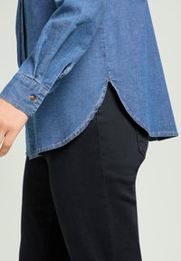 Niebieska dżinsowa koszula z długimi rękawami, z zaokrąglonym dołem, z zapięciem na guziki przy mankietach i lekko spłowiałą teksturą. Noszona na ciemnych spodniach.