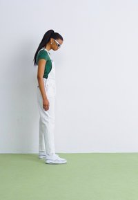 Hvite denimstøvler kombinert med en grønn skjorte, supplert med hvite sneakers. Subjektet har på seg solbriller og har langt, rett hår.