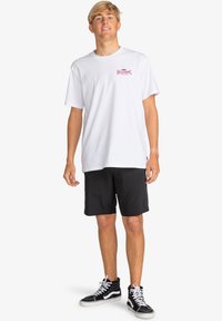 Camiseta de algodón blanca con un logo rosa en el pecho, combinada con pantalones cortos negros y zapatillas altas negras con acentos blancos.