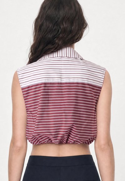 Femme aux longs cheveux foncés portant un top rayé court sans manches, bordeaux et blanc, montrant son dos et une partie d’un pantalon noir.
