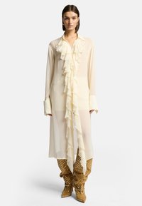 Blusa di crema chiara con dettagli a volant lungo la parte frontale, maniche lunghe e una vestibilità rilassata, abbinata a stivali al ginocchio marroni con pois.