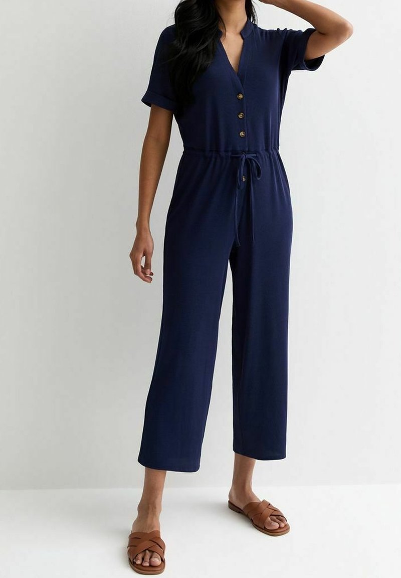 Femme portant une combinaison bleu marine boutonnée avec une taille à cordon et des jambes larges raccourcies, associée à des sandales plates marron.