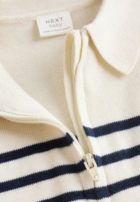 Gros plan sur un pull bébé en tricot crème avec des rayures horizontales bleu marine, fermeture éclair partiellement ouverte, et un col plié avec une étiquette "NEXT baby".