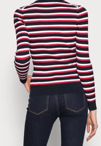 Maglione a coste in navy, rosso e bianco, con un design aderente e maniche lunghe. Abbinato a jeans in denim scuro.