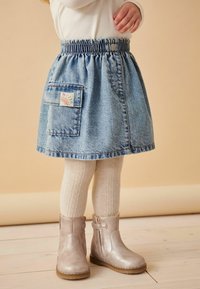 Jeansrock mit elastischem Bund, Vordertasche mit einem bunten "Hallo"-Patch. Kombiniert mit cremefarbenen Strumpfhosen aus Kabelstrick und silbernen Stiefeln.