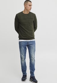 Olive green long-sleeve shirt with a chest pocket, paired with light blue denim jeans and black athletic shoes. Casual, relaxed fit.