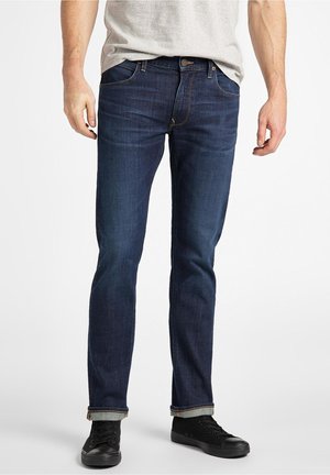 Homme portant un jean slim bleu foncé, un t-shirt gris clair et des baskets noires, debout devant un fond blanc.