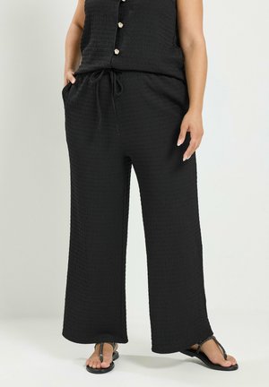 Vrouw draagt losse zwarte gestructureerde broek met voorzakken en een bijpassende mouwloze top, gecombineerd met zwarte teenslippers.