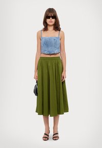 Top scurt din denim cu bretele subțiri, asortat cu o fustă maxi plisată verde. Modelul ține o geantă mică neagră și poartă sandale negre cu bretele.