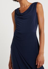 Vestido azul marino con escote drapeado, diseño sin mangas, textura suave y detalle fruncido en la cintura, creando una silueta fluida.