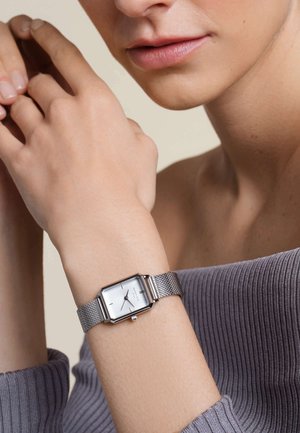 Montre rectangulaire argentée au poignet, dotée d'un bracelet en maille, d'un cadran épuré avec des marqueurs horaires minimalistes et d'une finition lisse. Haut gris doux.