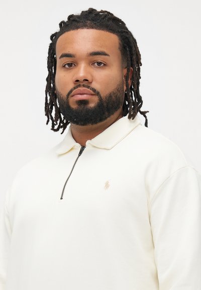 Άνδρας με γενειάδα και κοτσιδάκια που φοράει ένα κρεμ καφέ φανελάκι με φερμουάρ, έχοντας ένα μικρό κεντημένο λογότυπο στο στήθος, σε λευκό φόντο.