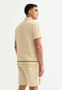 Beige short-sleeve shirt with textured fabric, featuring black floral patterns at the hem. Paired with matching beige shorts.