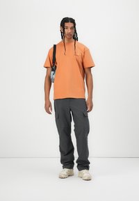 Oranje katoenen t-shirt met een klein logo, gecombineerd met grijze cargobroeken met meerdere zakken en beige sneakers.