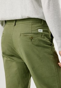 Olive green, cotton chino pants featuring a smooth texture, buttoned back pocket, and a small brand label near the waistband.