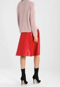 Kvinna iklädd en blek rosa tröja, röd knälång läderskjorta och svarta högklackade ankelstövlar, står med ryggen vänd.