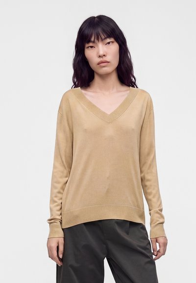 Femme aux longs cheveux noirs portant un pull beige à col en V et un pantalon noir ample, debout devant un fond clair uni.