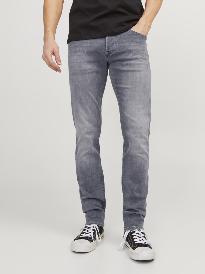 Eng anliegende graue Jeans aus Denim, mit glatter Textur und Fünf-Taschen-Design. Kombiniert mit schwarzen Sneakers mit weißen Schnürsenkeln.