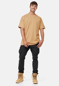 Camiseta de algodón beige de manga corta, combinada con pantalones cargo negros con bolsillos laterales y botas de cordones marrones con suela gruesa.