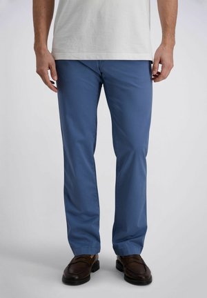 Mann trägt blaue gerade geschnittene Hose, weißes Hemd und dunkelbraune Lederschuhe, steht vor schlichtem Hintergrund.