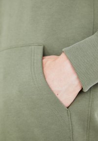 Felpa verde oliva con polsini a costine texture e tasca laterale. Il materiale sembra morbido, con dettagli di cucitura visibili.