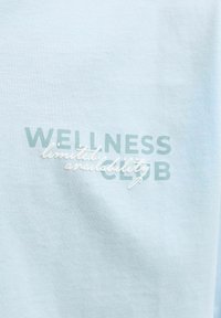Ανοιχτό μπλε ύφασμα με το κείμενο «WELLNESS CLUB» σε πράσινα γράμματα κεφαλαία και «περιορισμένη διαθεσιμότητα» με λευκή πλάγια γραφή που επικαλύπτει το κείμενο.