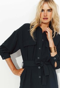 Robe noire avec col, boutonnière à l'avant et deux poches poitrine. Possède une taille ceinturée et des manches retroussées, fabriquée dans un tissu lisse.