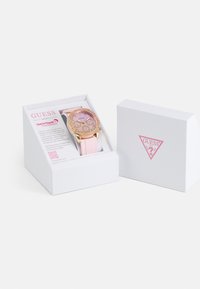 Montre avec bracelet en silicone rose et boîtier en acier inoxydable doré rose, dotée d'un cadran orné. Présentée dans une boîte blanche avec un logo rose et une carte de détails du produit.