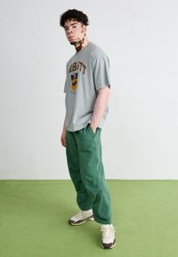 Grå oversized t-shirt med svart och orange grafik, tillsammans med gröna baggy byxor och vita sneakers med bruna accenter på en grön bakgrund.
