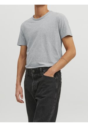 Hombre con una camiseta de manga corta gris claro metida dentro de unos pantalones vaqueros negros de cintura alta, con una mano en el bolsillo y un tatuaje delgado en el brazo.