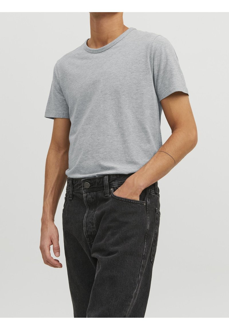 Homme portant un t-shirt à manches courtes gris clair rentré dans un jean noir taille haute, avec une main dans sa poche et un mince tatouage sur le bras.