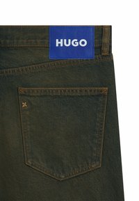 Jeans de denim oscuro con un parche de cuero azul etiquetado "HUGO." Presenta un bolsillo trasero y detalles de costura visibles.