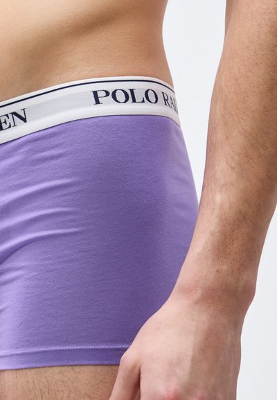 Πλευρική όψη ατόμου που φοράει ανοιχτό μοβ ανδρικά εσώρουχα με λευκή ζώνη μέσης που εμφανίζει το κείμενο "Polo Ralph Lauren".